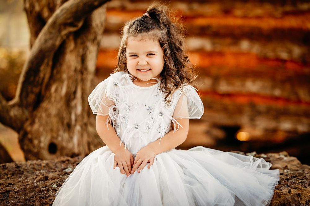 A child in a white lace dress sits outdoors during a portrait session with rustic wooden backgrounds.