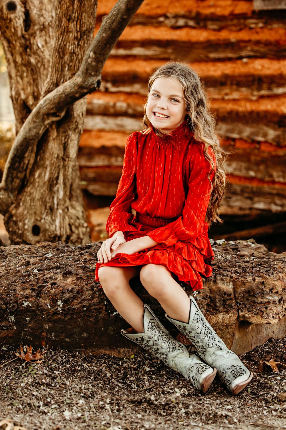 A red dress and silver boots create a charming rustic portrait against a wooden log cabin backdrop.