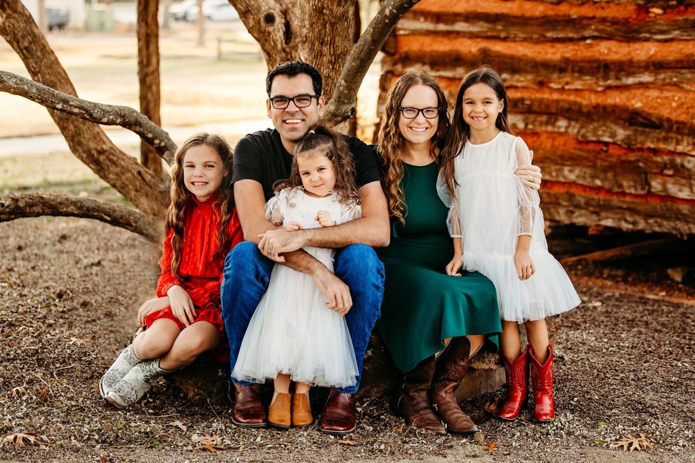 A family sits together outdoors against a rustic wooden wall in coordinated casual outfits.