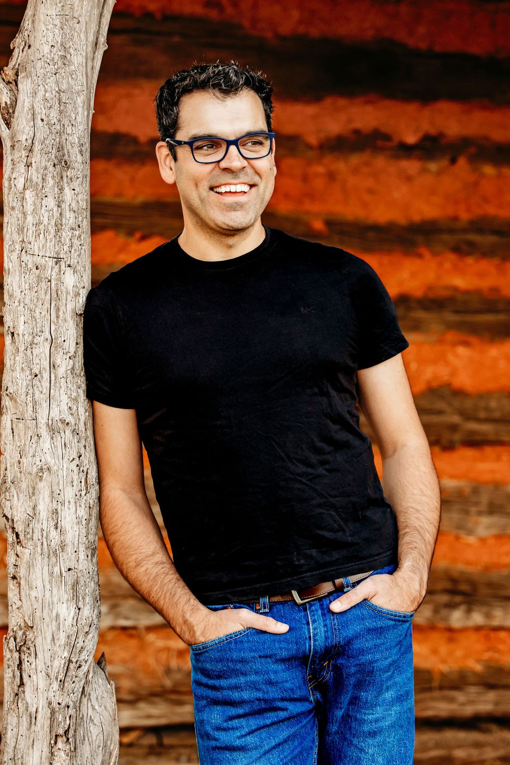 Person wearing a black t-shirt and blue jeans poses against a rustic wooden wall with dramatic orange lighting.