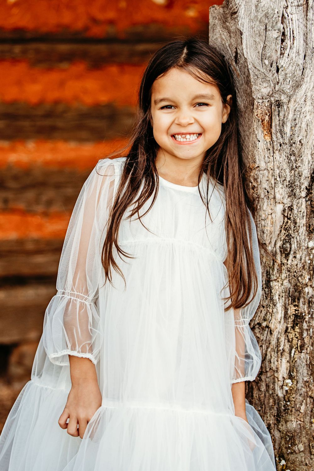 Young child in a white dress with sheer sleeves poses against rustic orange wooden wall and tree trunk.