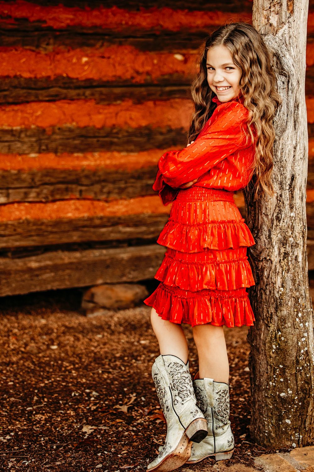 A person in a bright red ruffled dress and white cowboy boots poses against rustic wooden walls in autumn.