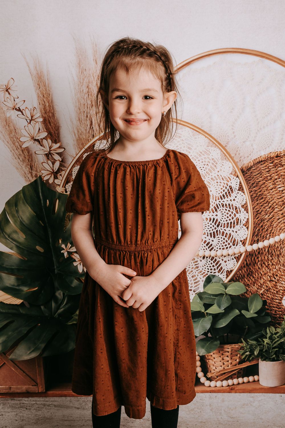 Young child in a brown dress poses against a bohemian backdrop with plants and woven decor.