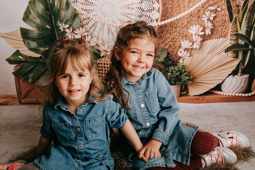Two young children wearing matching denim outfits pose against a boho backdrop with palm leaves and macrame.