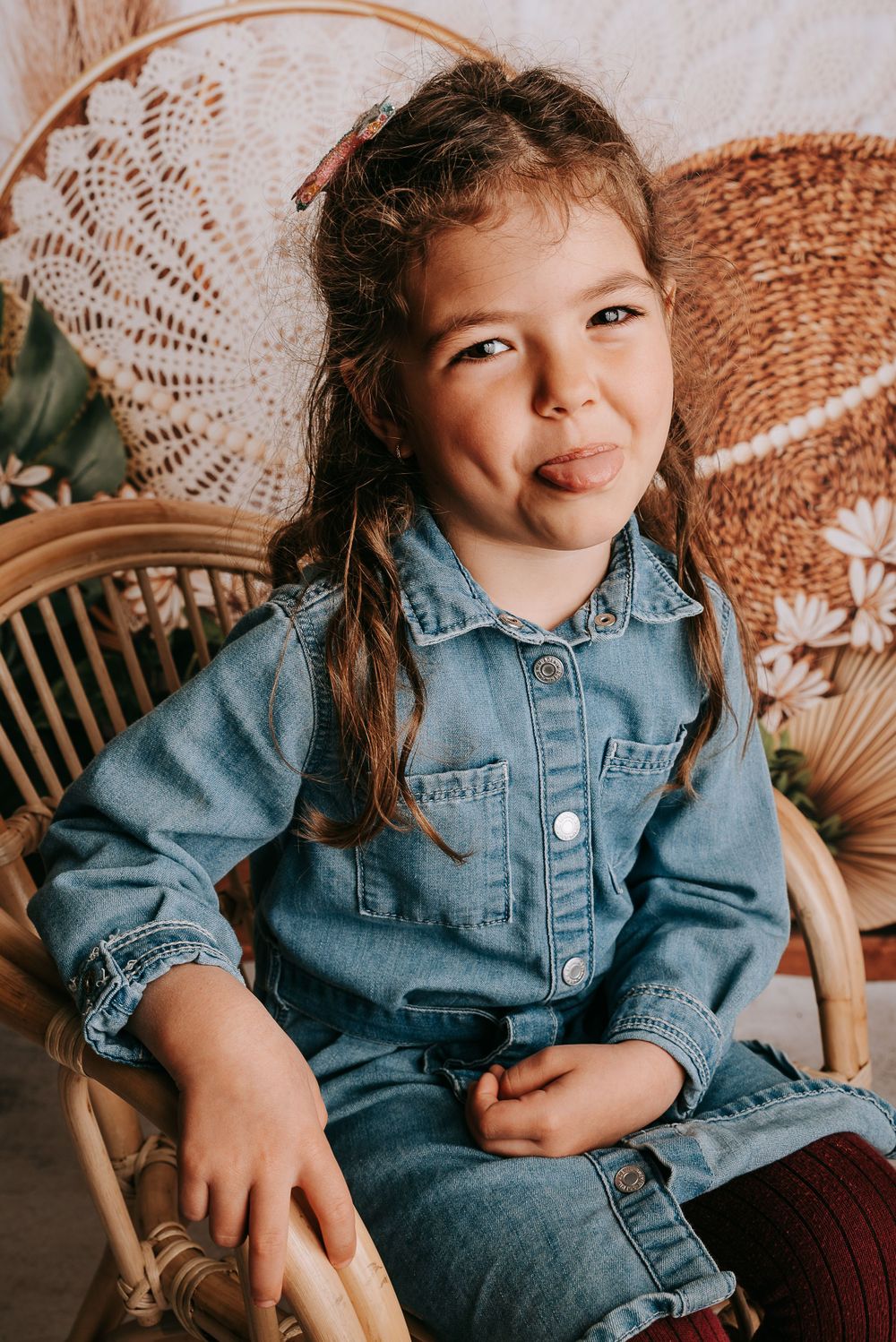 Playful portrait of child in denim outfit sitting on rattan chair with boho decorations.