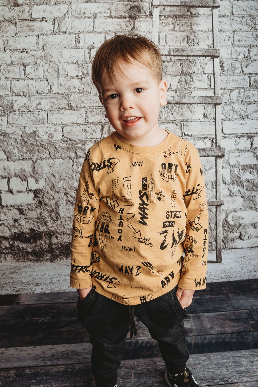 Child wearing tan graphic print sweater poses against white brick wall background.