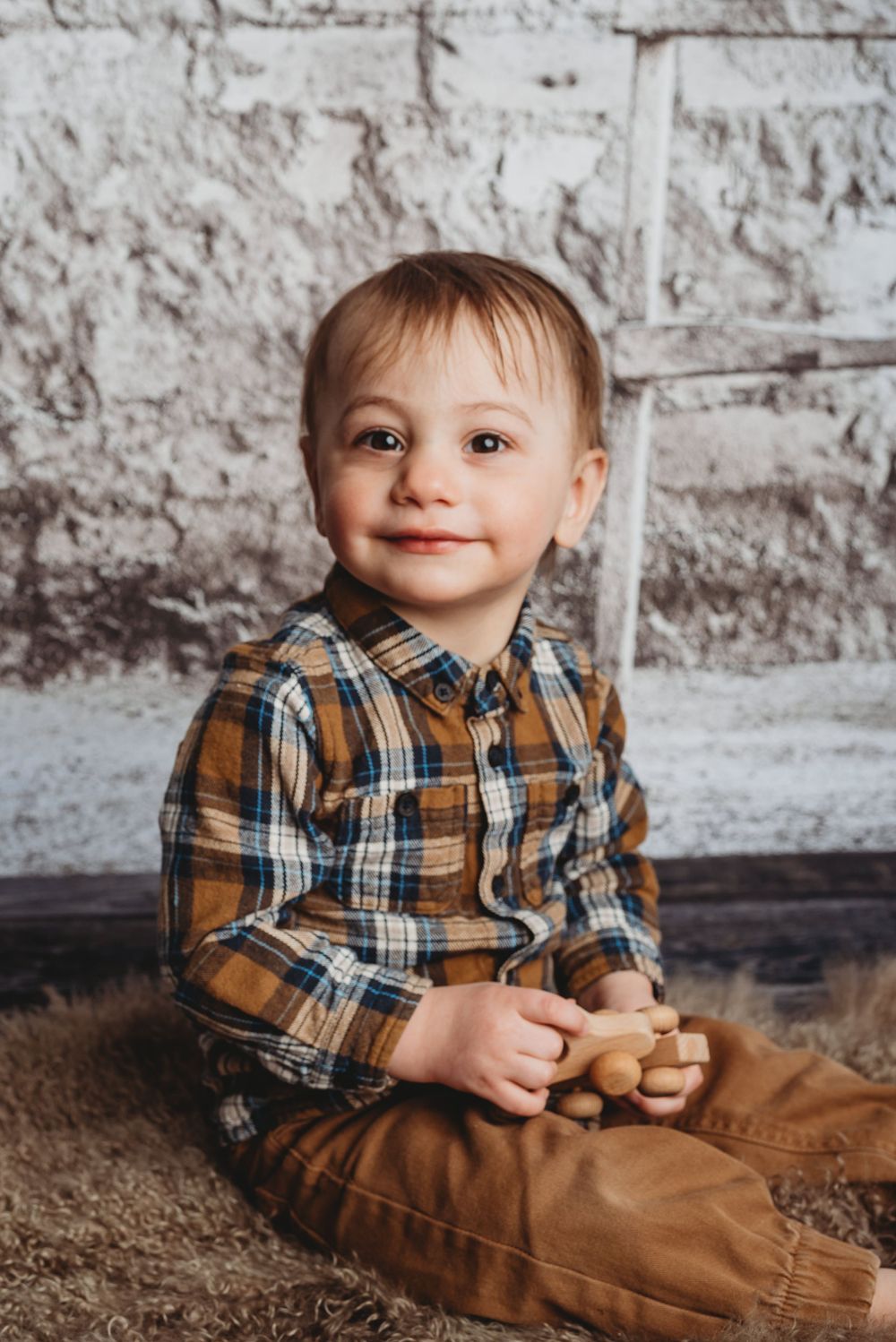 Child in plaid shirt sits on brown carpet against white brick wall background.