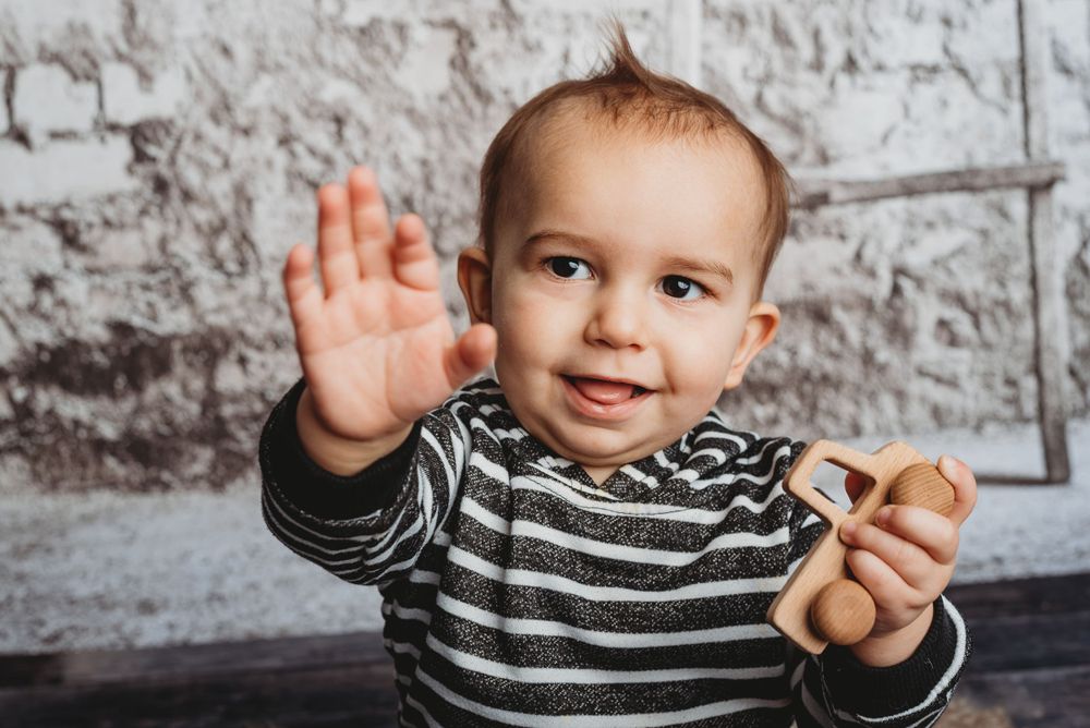 Candid shot of young child in striped shirt waving at camera against brick wall.