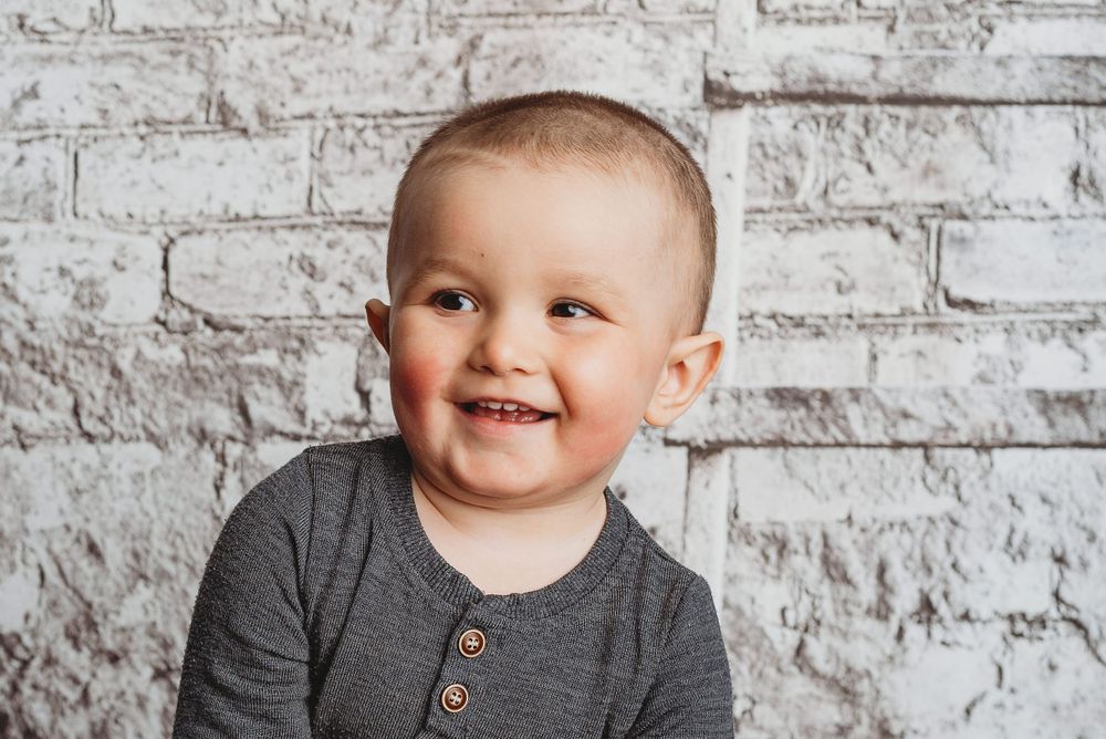 Joyful portrait of young child in gray henley shirt against white brick wall background.