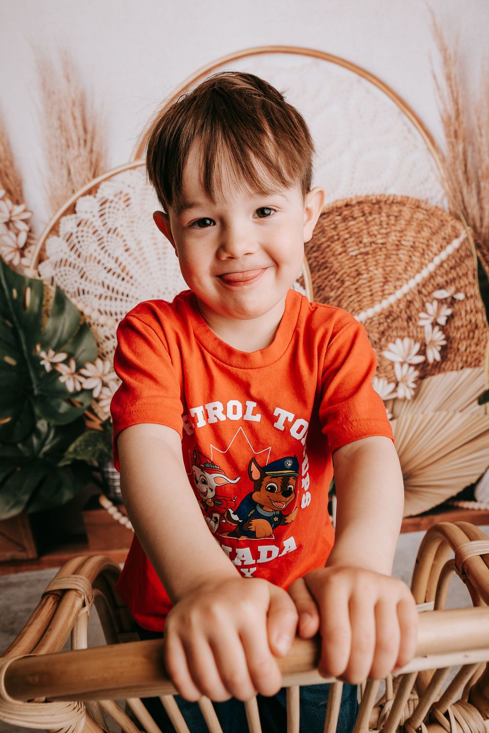 Smiling child in orange Paw Patrol shirt posing against woven backdrop with dried pampas grass.