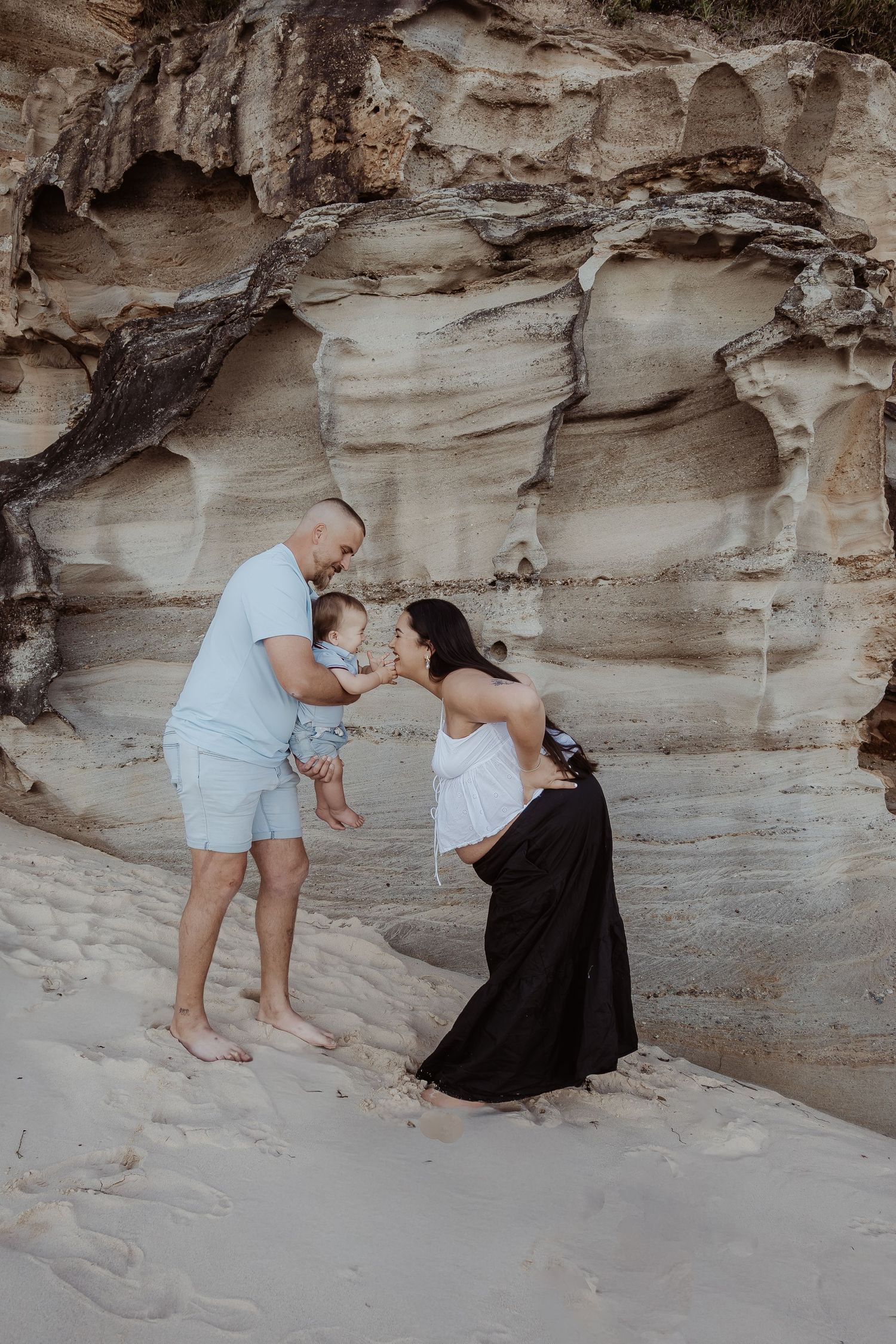 A family share a tender moment together against a backdrop of sandstone cliffs at the beach during a central coast maternity photoshoot.