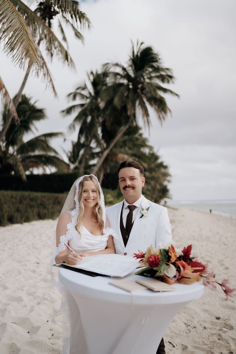 A couple in wedding attire sits on a white couch at a tropical beach with palm trees and a bouquet.