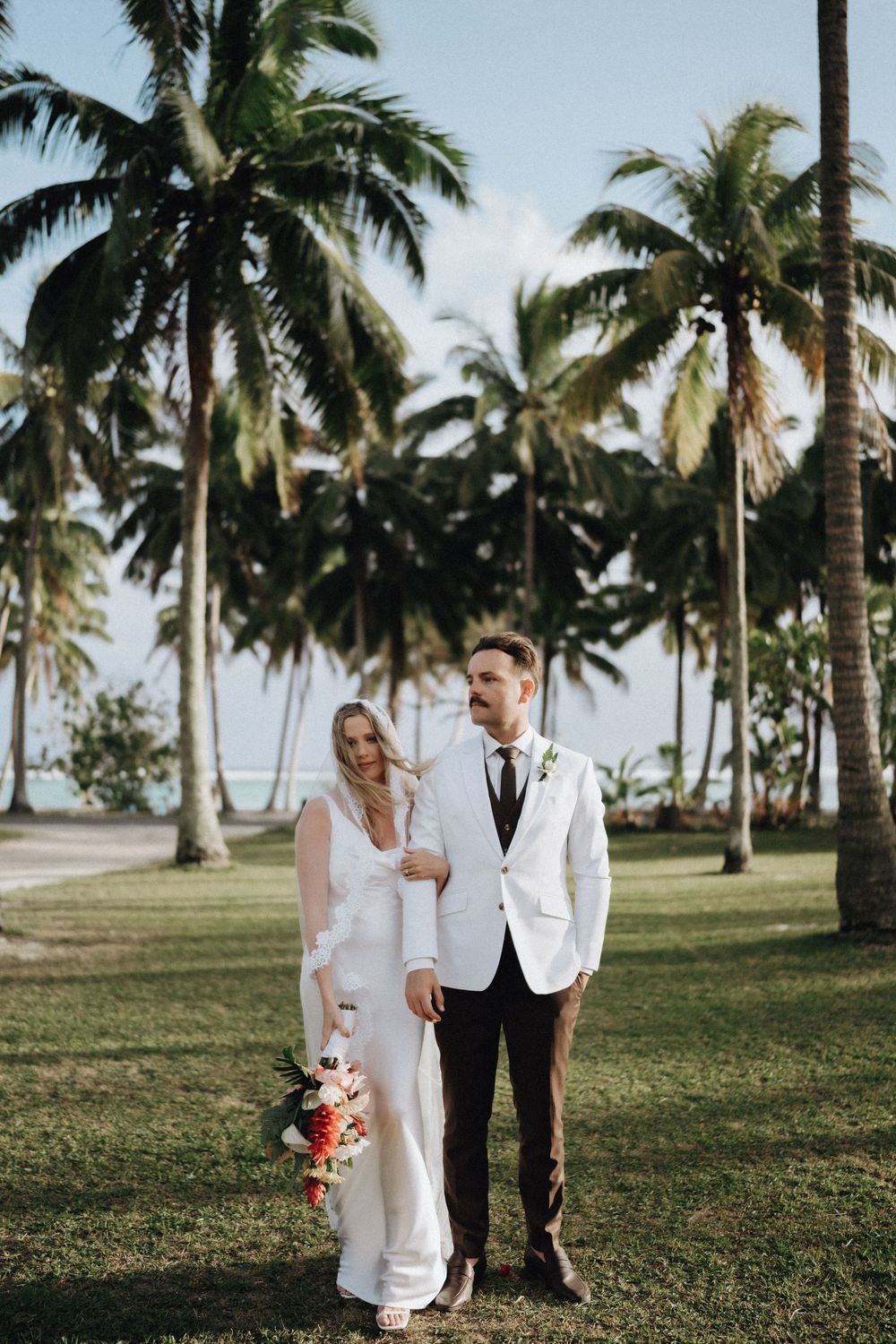 A couple in wedding attire shares a romantic moment among palm trees on a grassy tropical beach setting.