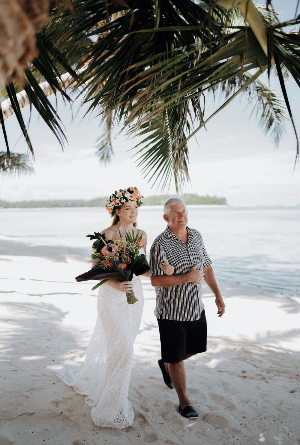 A couple runs playfully along a tropical beach with palm trees and white sand as waves crash behind them.