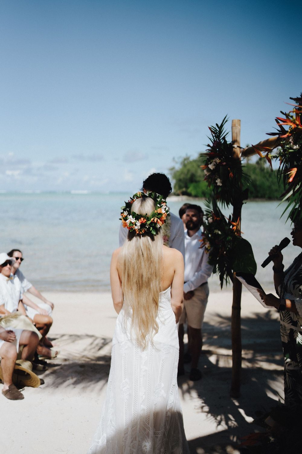 A beach wedding ceremony in progress with tropical decor and guests gathered on a sandy shoreline.