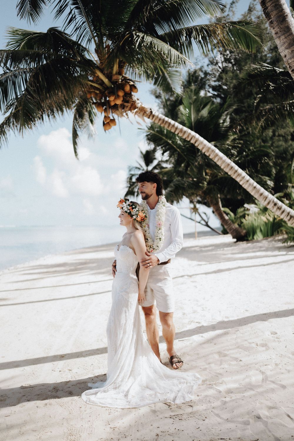 A romantic couple in white attire embraces on a tropical beach with palm trees swaying in the background.