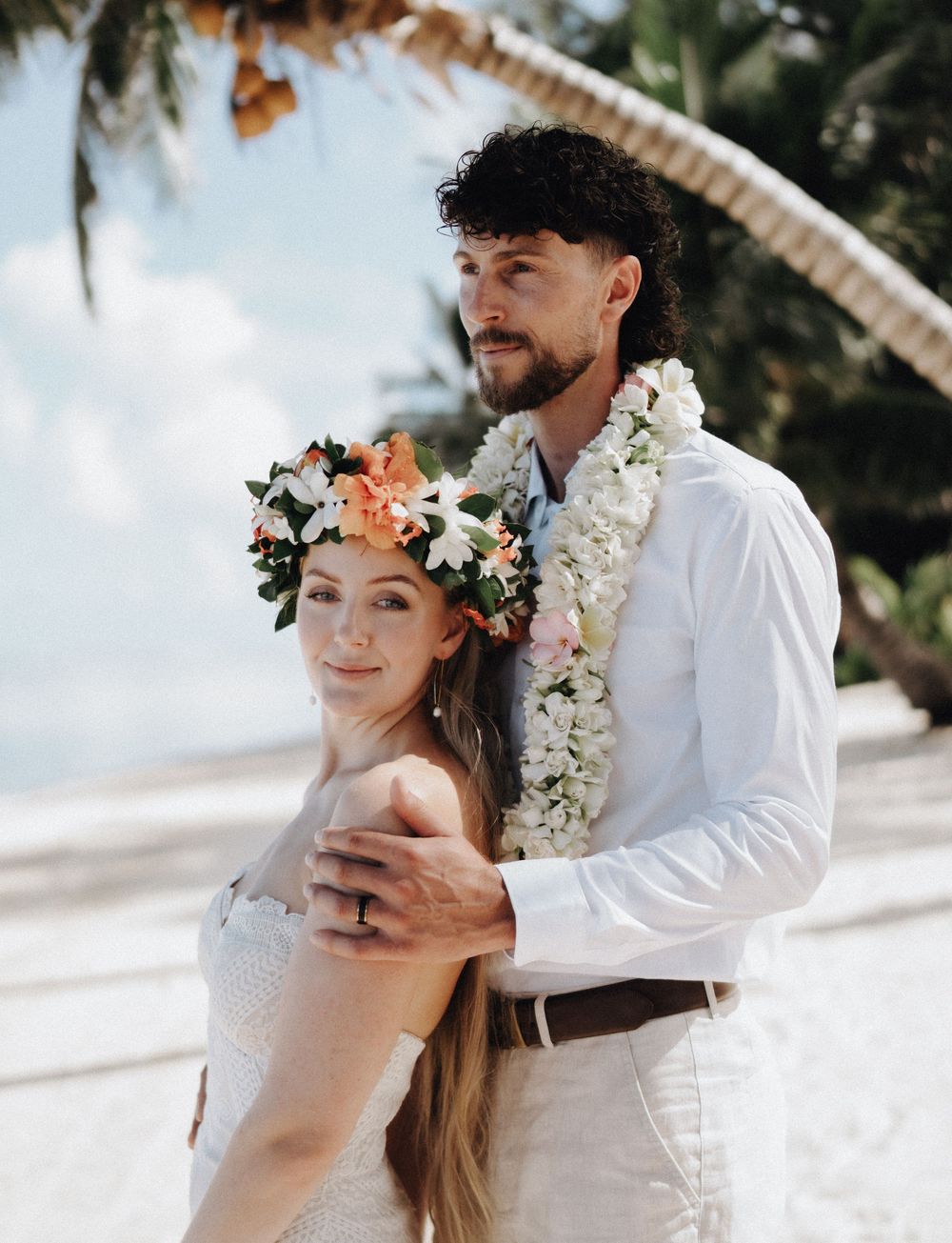 A couple in white attire shares intimate moments during a beach wedding photoshoot with floral accessories.