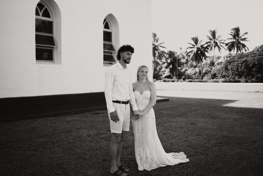 A couple poses together on a lawn outside a white church with arched windows and palm trees in the background.