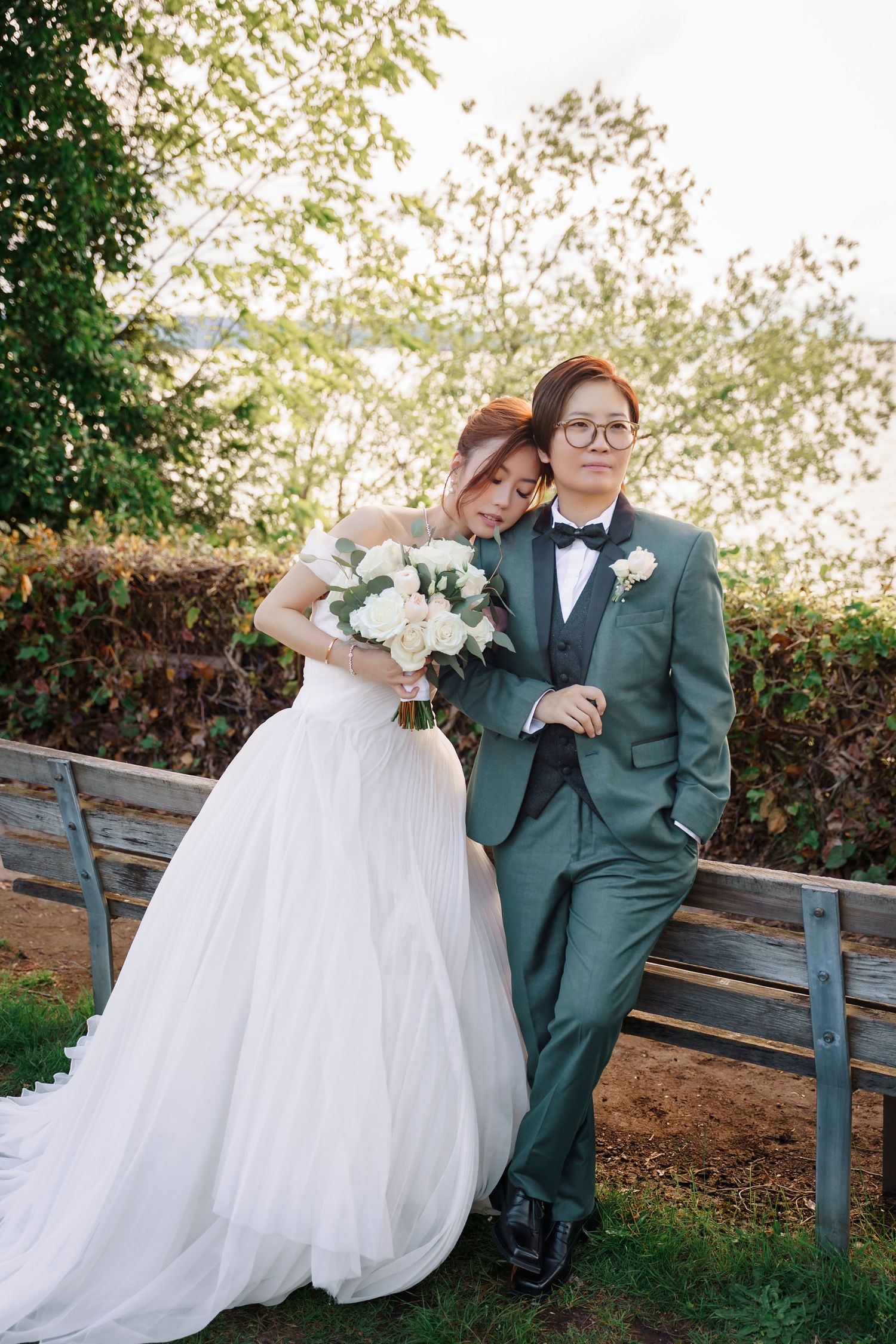 A romantic wedding couple sits together on a wooden bench in an outdoor garden setting.