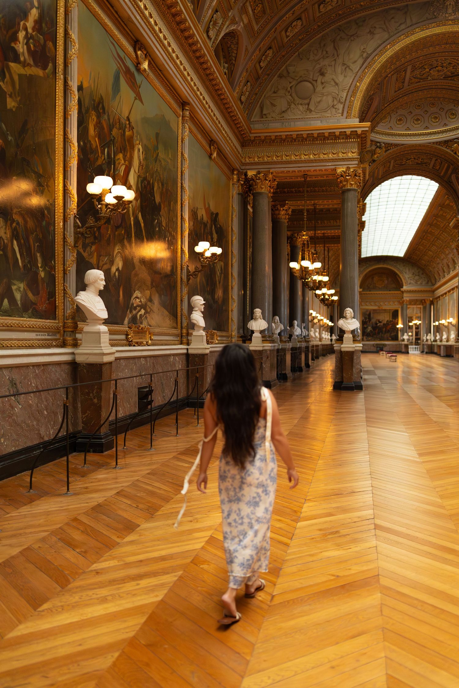 A long ornate gallery hall with polished wooden floors, gilded walls, and classical busts displayed along its length.
