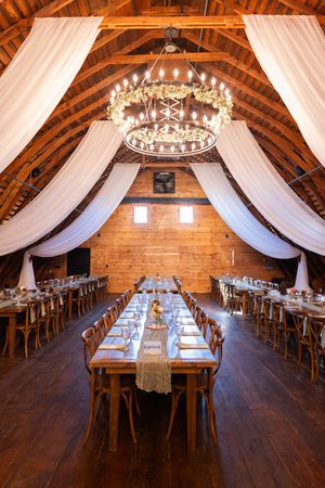 Rustic barn reception venue with wooden tables, white draping, and elegant chandeliers in a vaulted ceiling space.