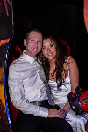 A couple in matching white attire smiles together at a dimly lit event with colorful lighting in the background.