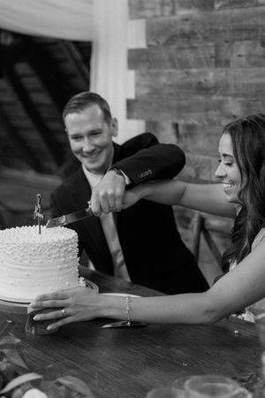 A couple laughs together while sharing a white frosted cake at a wooden table in black and white photography.