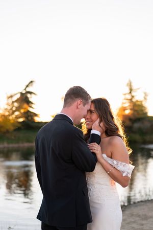 A romantic couple embraces during golden hour sunset by a lake, with the bride in white and groom in black suit.