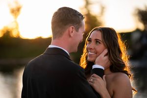 A romantic couple in formal attire embraces during sunset at their outdoor wedding celebration.