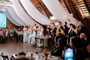 Wedding guests raise their glasses in celebration during a toast at a rustic venue decorated with white draping.