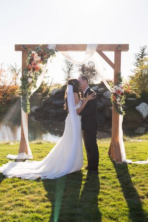 Silhouettes of a wedding couple standing under a wooden arch during golden hour sunset ceremony.