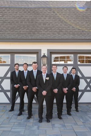 A row of groomsmen in black suits and ties standing against a white building exterior.