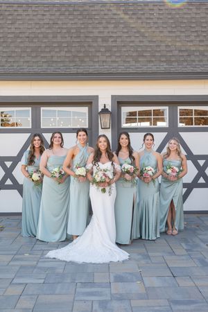 Wedding party in dusty blue bridesmaid dresses poses with bride in white gown on stone patio against gray wall.