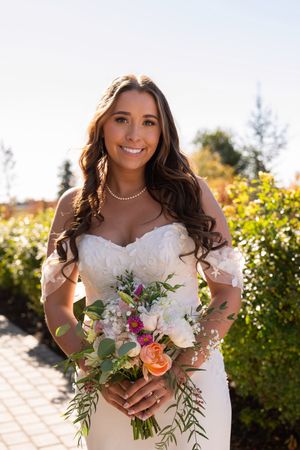 Bride in white lace gown holding colorful bouquet with roses and greenery in sunny garden setting.
