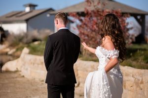 A couple in wedding attire stands together outdoors, with the bride in white and groom in black against a scenic backdrop.