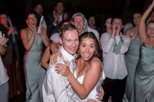 A happy couple dances together at their wedding reception while guests in sage green dresses celebrate in the background.
