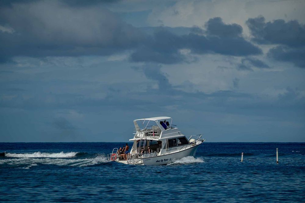 A series of boats cruise across blue ocean waters under cloudy skies near a tropical destination.