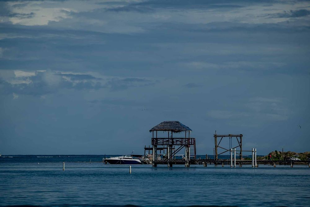 A wooden dock structure with thatched roof stands over calm blue waters against a cloudy sky in the Caribbean.