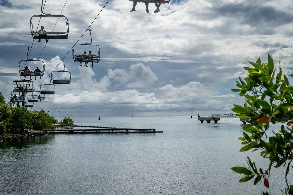 Ski lift chairs suspended over a tranquil lake with lush greenery and dramatic cloud formations.