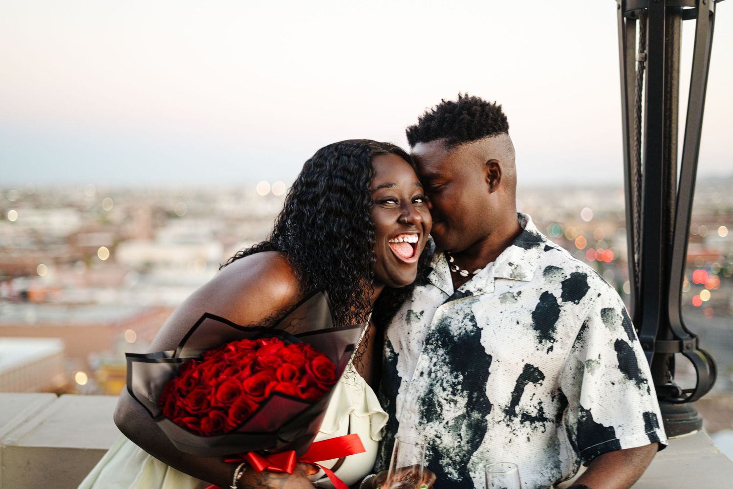 A romantic couple embraces against a scenic city backdrop during what appears to be an engagement shoot.