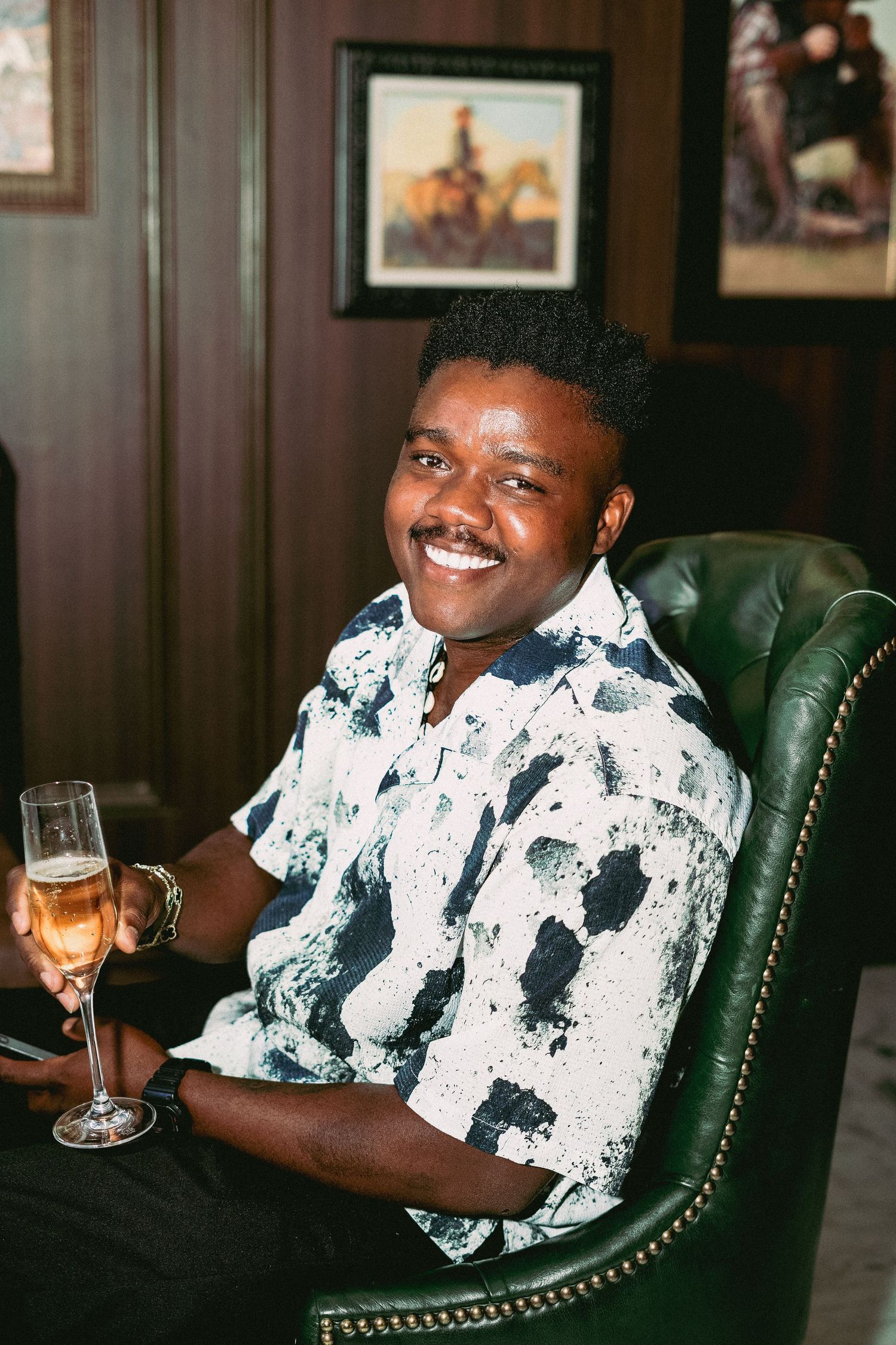 A person in a black and white patterned shirt sits in a green leather chair holding a wine glass.