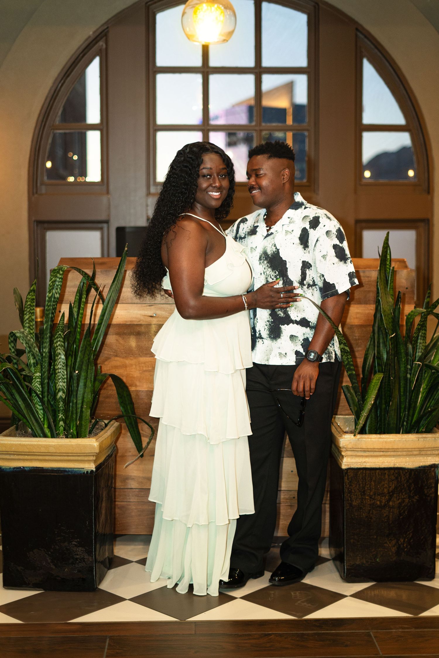 A couple shares a joyful moment together in an elegant indoor venue with arched windows and decorative plants.