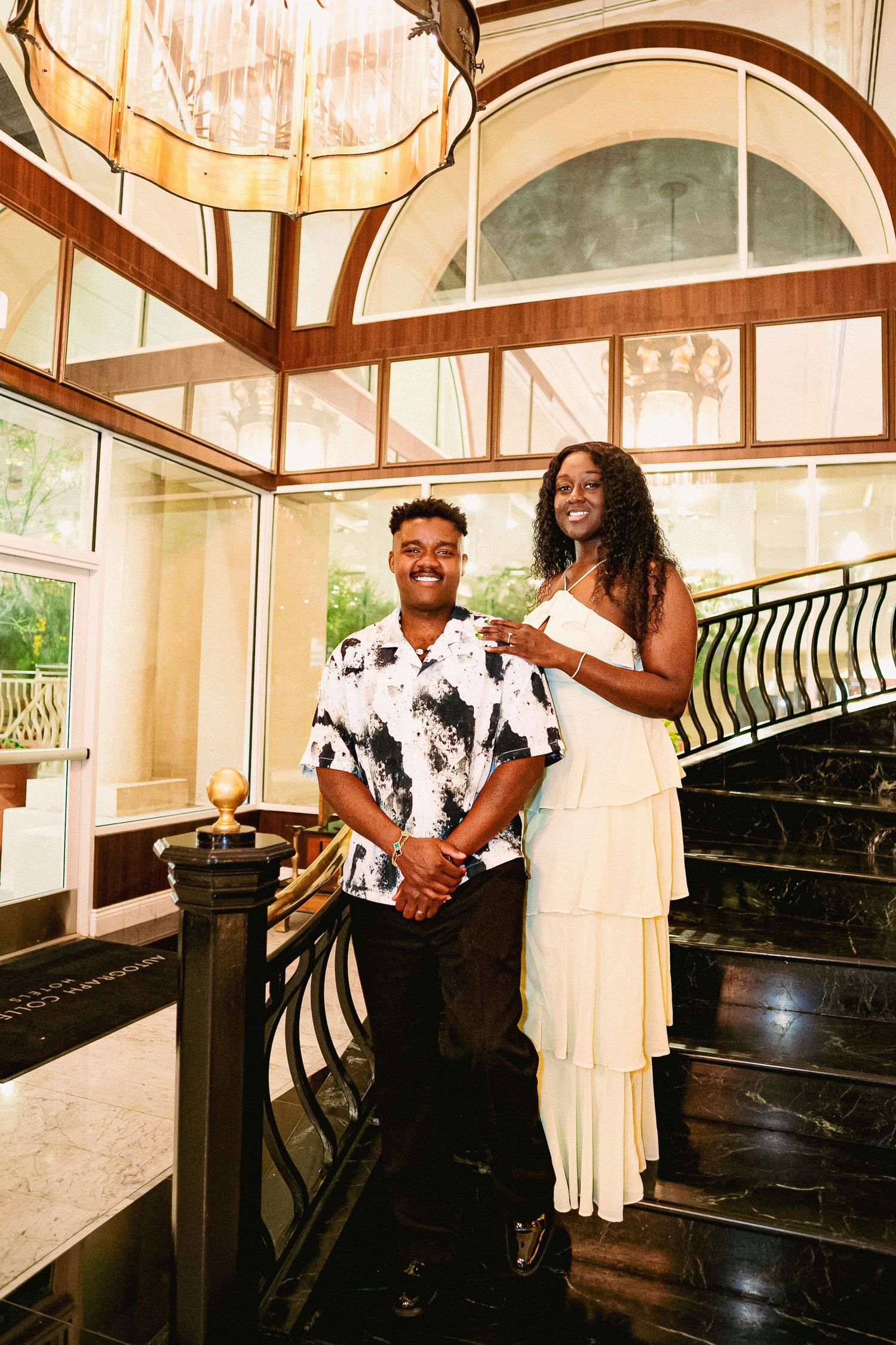 A couple poses together in an elegant indoor setting with a piano and curved staircase in warm lighting.