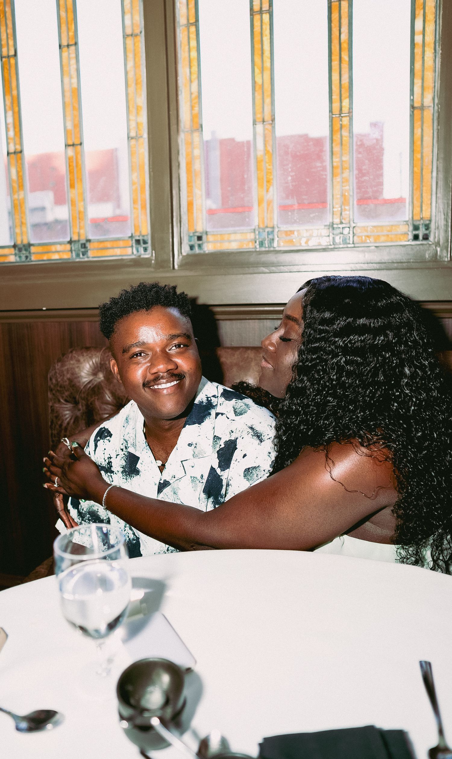 Two people share a joyful moment while dining at a restaurant with tall windows and city views in the background.