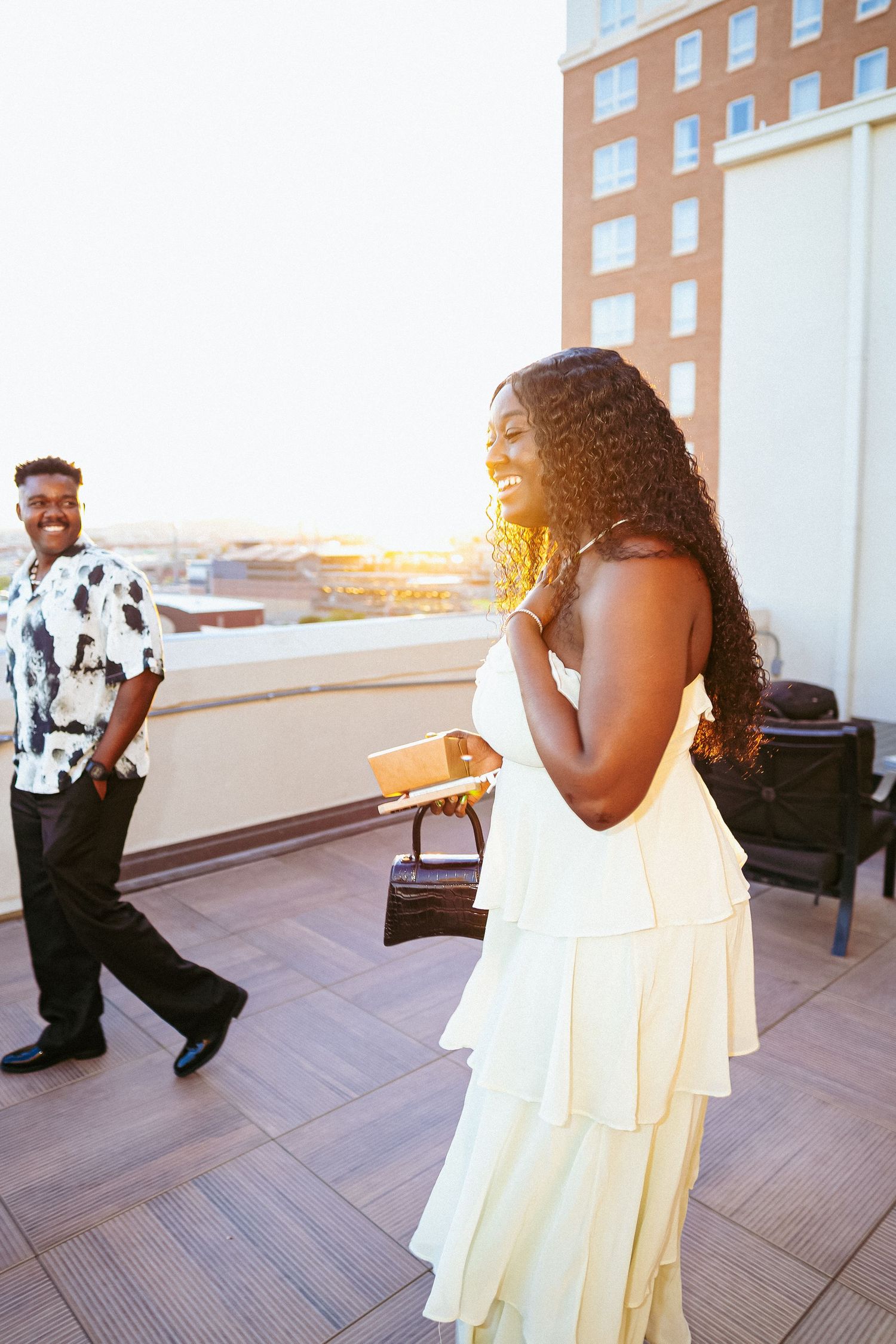 A couple shares a romantic moment on a rooftop terrace at sunset with a brick building in the background.