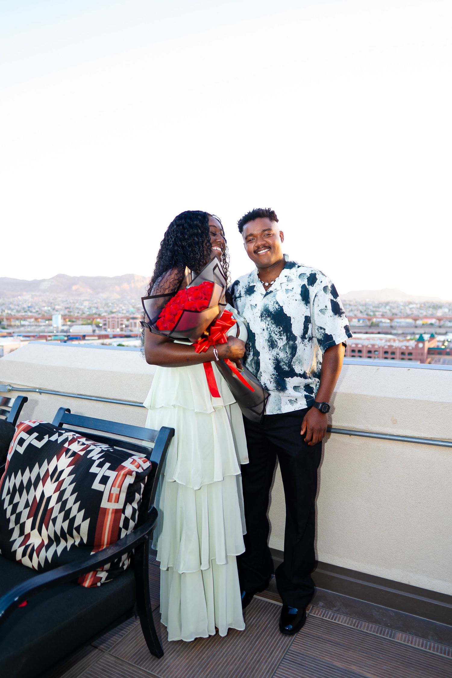 A romantic couple in black and white attire embrace on a rooftop terrace with city views in the background.