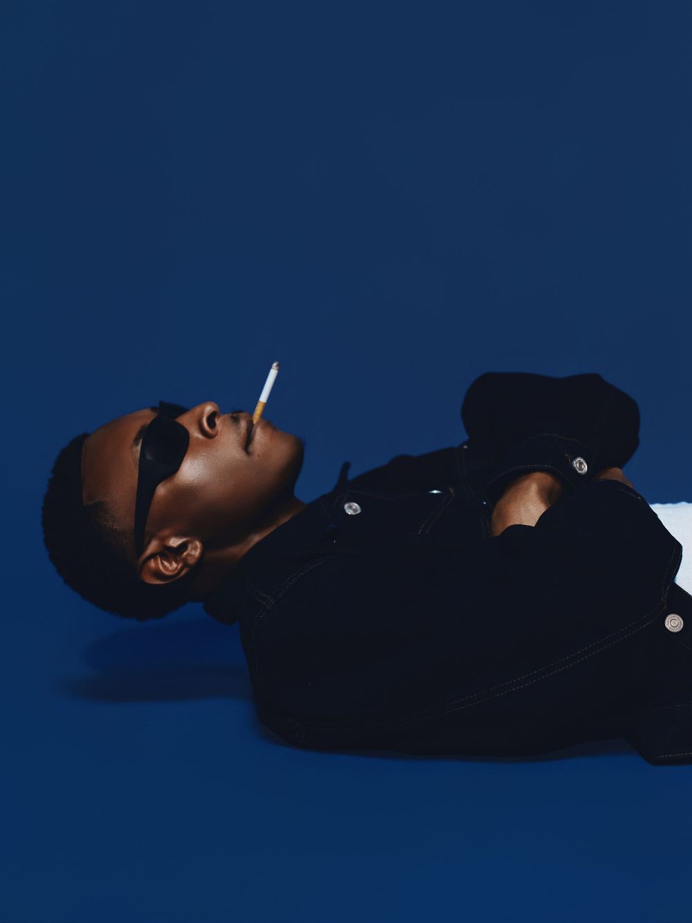 A person wearing a black outfit lies on their back against a dark blue background in a dramatic pose.