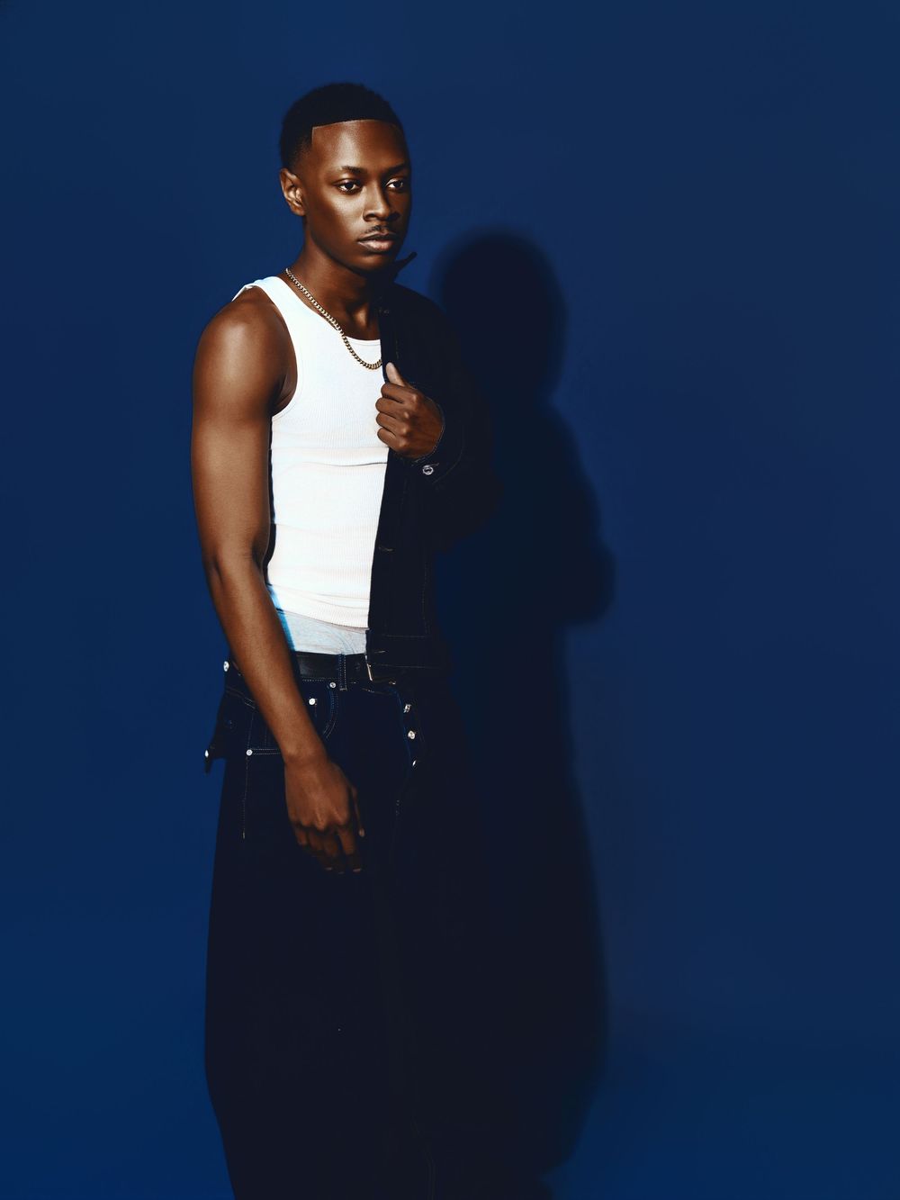 Person in a white tank top and dark pants poses dramatically against a deep blue background with strong shadows.