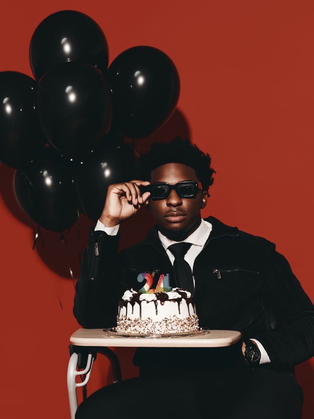 Stylish figure in black suit and hat poses with black balloons against red background for dramatic party photoshoot.