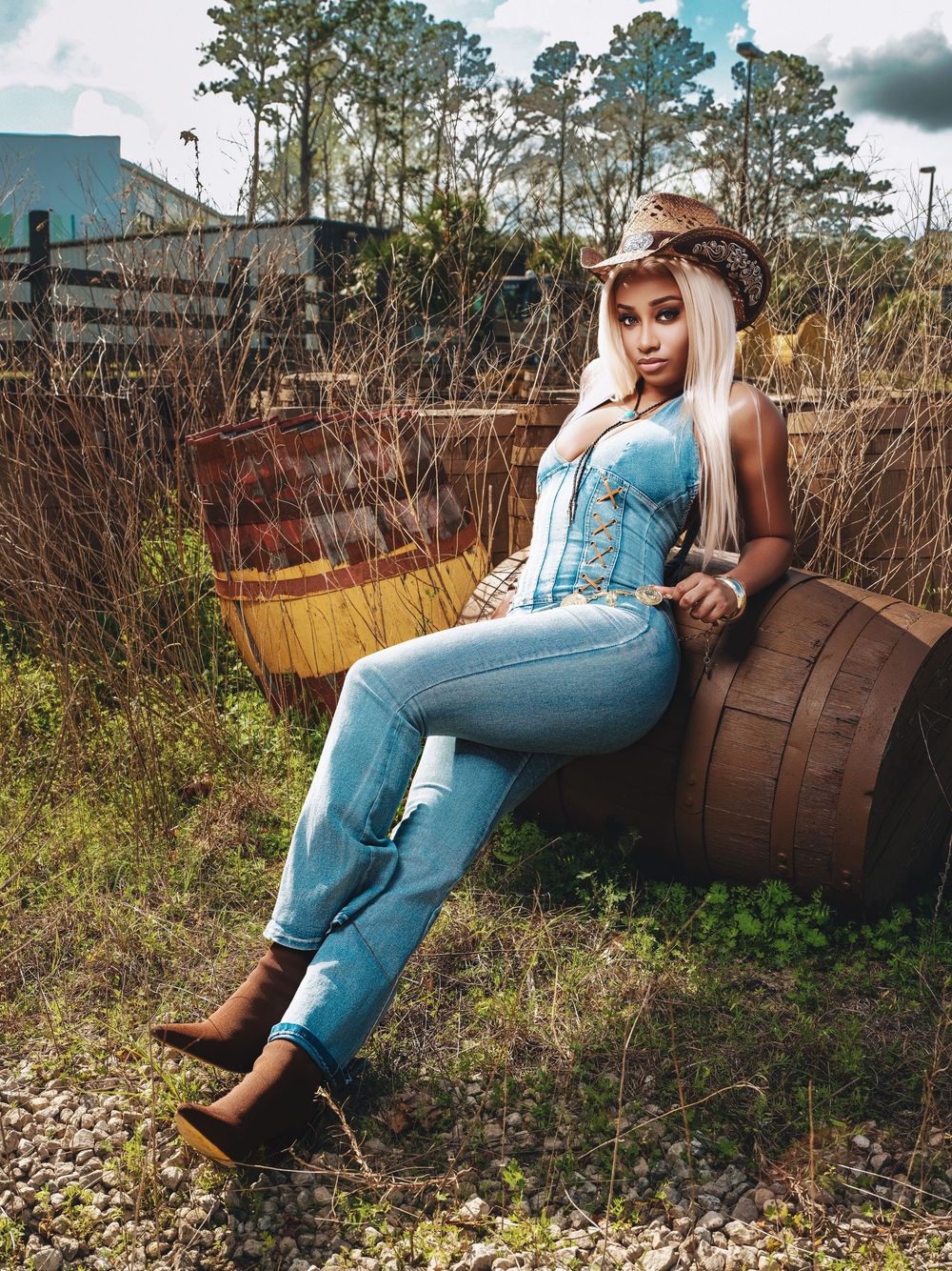 Country western styled model in denim and corset poses on rustic wooden barrel in outdoor setting with pine trees.
